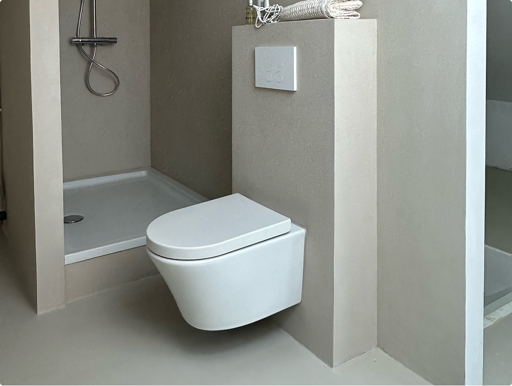 Gästebad von X²O Badezimmer mit weißem wandhängendem WC neben einer flachen Duschwanne, beigen Wänden und halbhoher Wand mit weißer Betätigungsplatte.