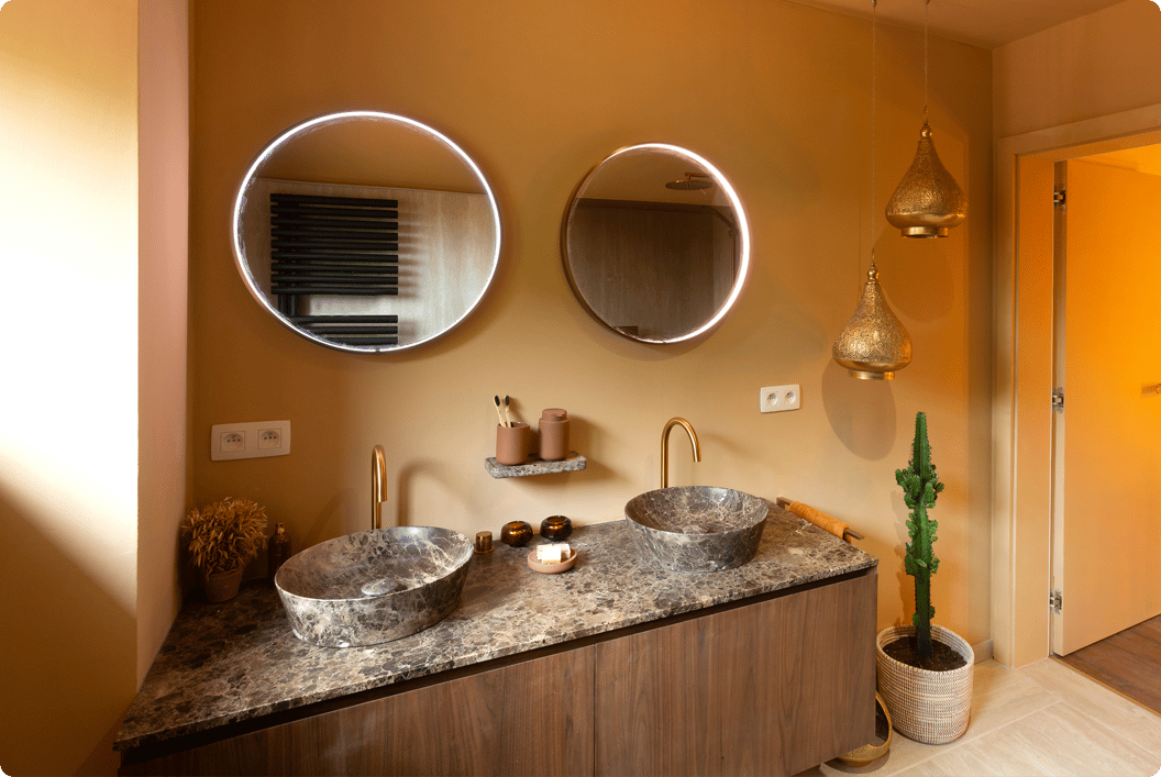 Ambiance orientale chaleureuse dans cette salle de bains x²o avec des tons terre, deux miroirs ronds à éclairage intégré et un meuble lavabo double en bois avec plan de travail effet marbre. Des lanternes suspendues et un cactus en pot tressé sont visibles près de la porte.