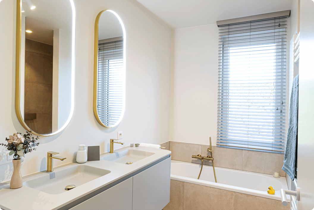 Salle de bains x²o lumineuse et fraîche avec une disposition moderne. À gauche, meuble suspendu avec double vasque, robinets dorés et deux miroirs verticaux à éclairage LED. À droite, une baignoire encastrée avec robinet assorti et une fenêtre avec des stores laisse entrer la lumière naturelle.