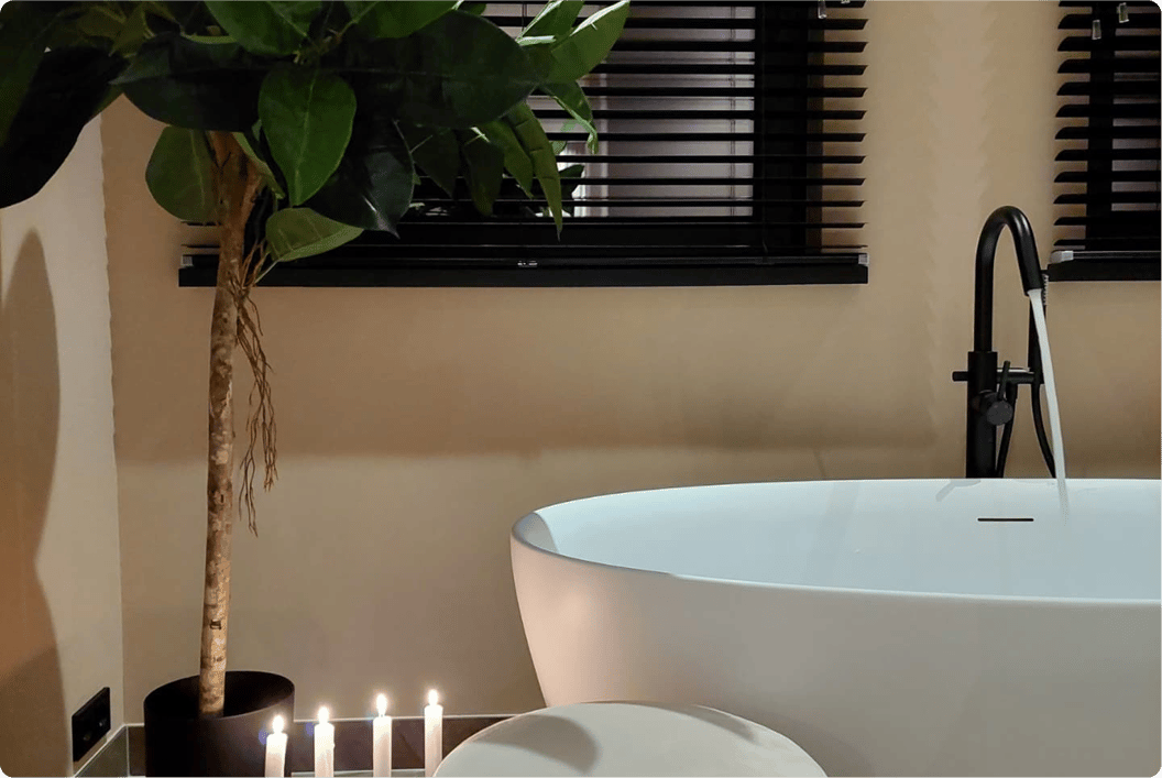 Salle de bains x²o chaleureuse avec baignoire blanche îlot, robinet noir sur pied et ambiance tamisée. Six bougies allumées et une grande plante verte ajoutent une touche relaxante. En arrière-plan, des stores noirs couvrent la fenêtre.