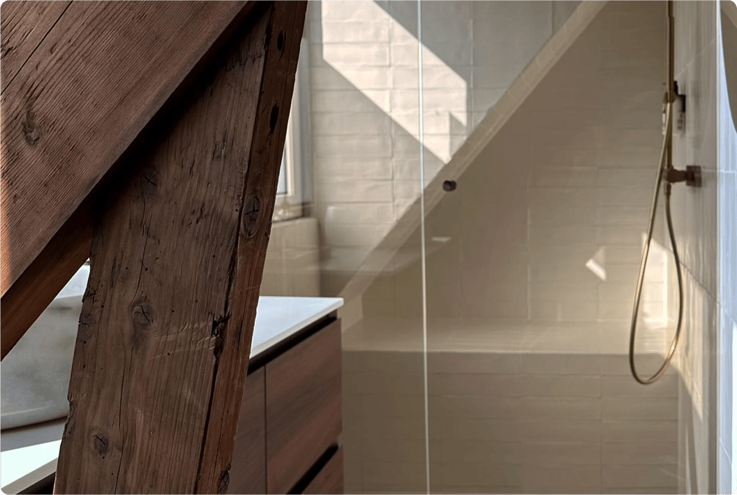 Salle de bains x²o pleine de caractère avec plafond incliné et poutres en bois apparentes. À droite, une douche avec carrelage blanc et pommeau pluie doré. À gauche, un meuble de salle de bains en bois sous une fenêtre laissant entrer la lumière du jour.
