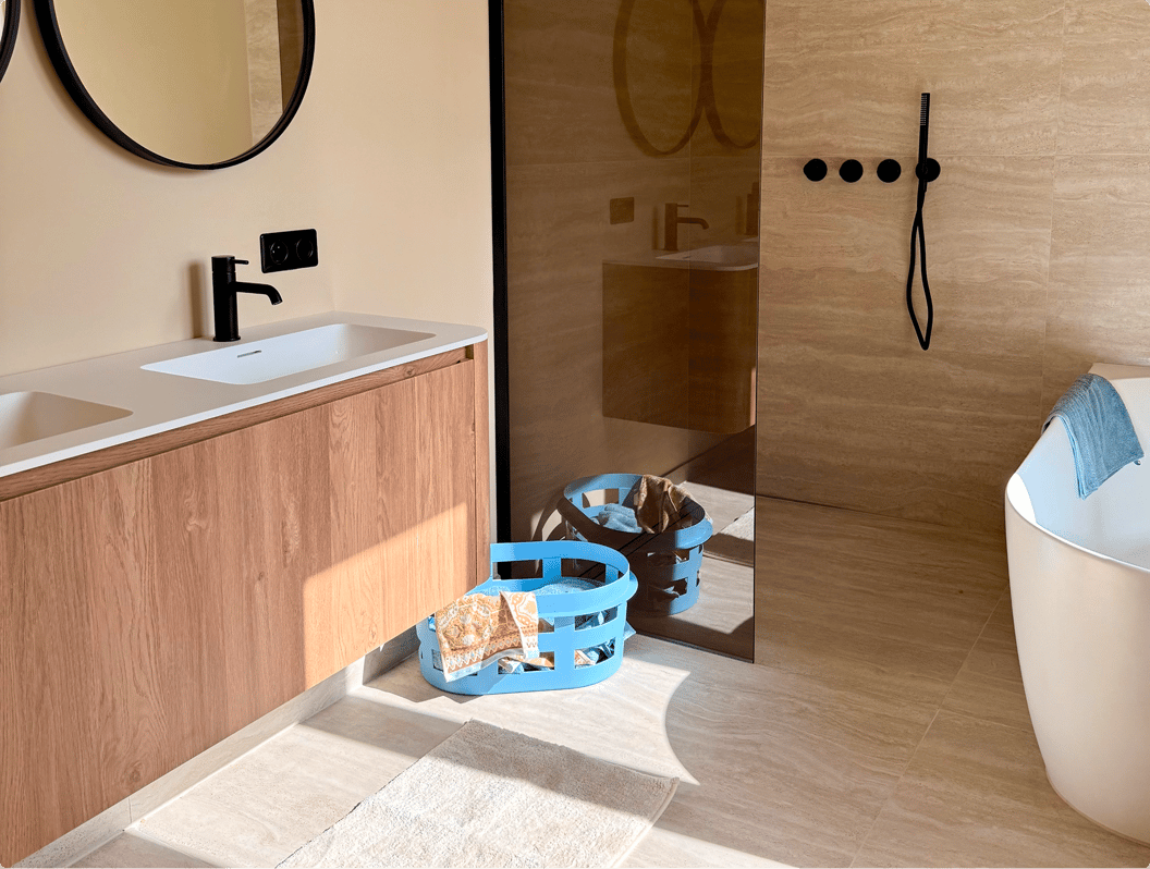 In diesem realistischen Badezimmer von x²o Badezimmer siehst du einen Waschtischunterschrank aus Holz mit integriertem weißen Waschbecken, eine begehbare Dusche mit getönter Glaswand und schwarzen Duscharmaturen, eine freistehende Badewanne mit Handtuch und einen blauen Wäschekorb auf einem hellen Fliesenboden.
