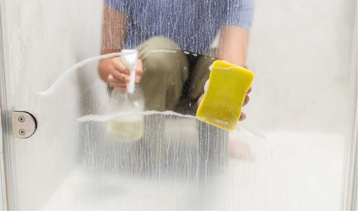 Reinigung einer nassen Duschwand mit gelbem Schwamm und Spray – X²O Badezimmer