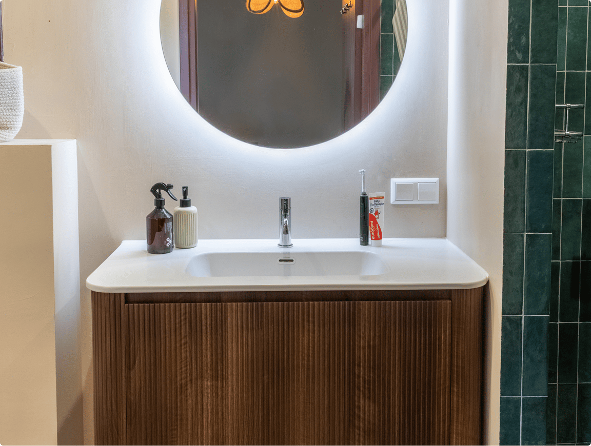 NL : Modern wastafelmeubel van x²o badkamers in geribbeld hout met geïntegreerde witte waskom, chromen kraan, accessoires zoals zeepdispenser en tandenborstel, afgewerkt met een ronde verlichte spiegel en rechts een douche met groene tegels en glazen wand