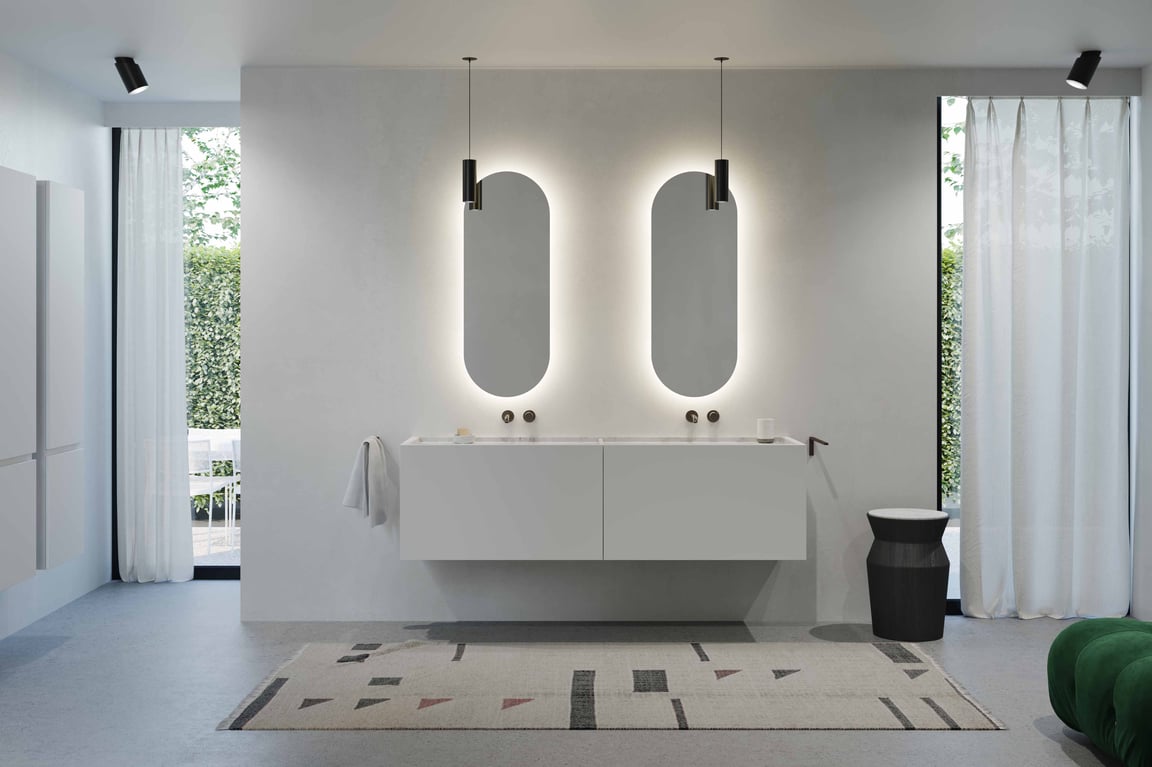 Minimalistische badkamer met zwevend wit badkamermeubel uit de Balmani Cubo collectie van x²o badkamers, afgewerkt in witte lak met een geïntegreerde Carrara marmeren wastafel. Boven het meubel hangen twee langwerpige, afgeronde spiegels met indirecte ledverlichting. De ruimte heeft lichte muren, een groot raam met witte gordijnen en een strak, rustig interieur.
