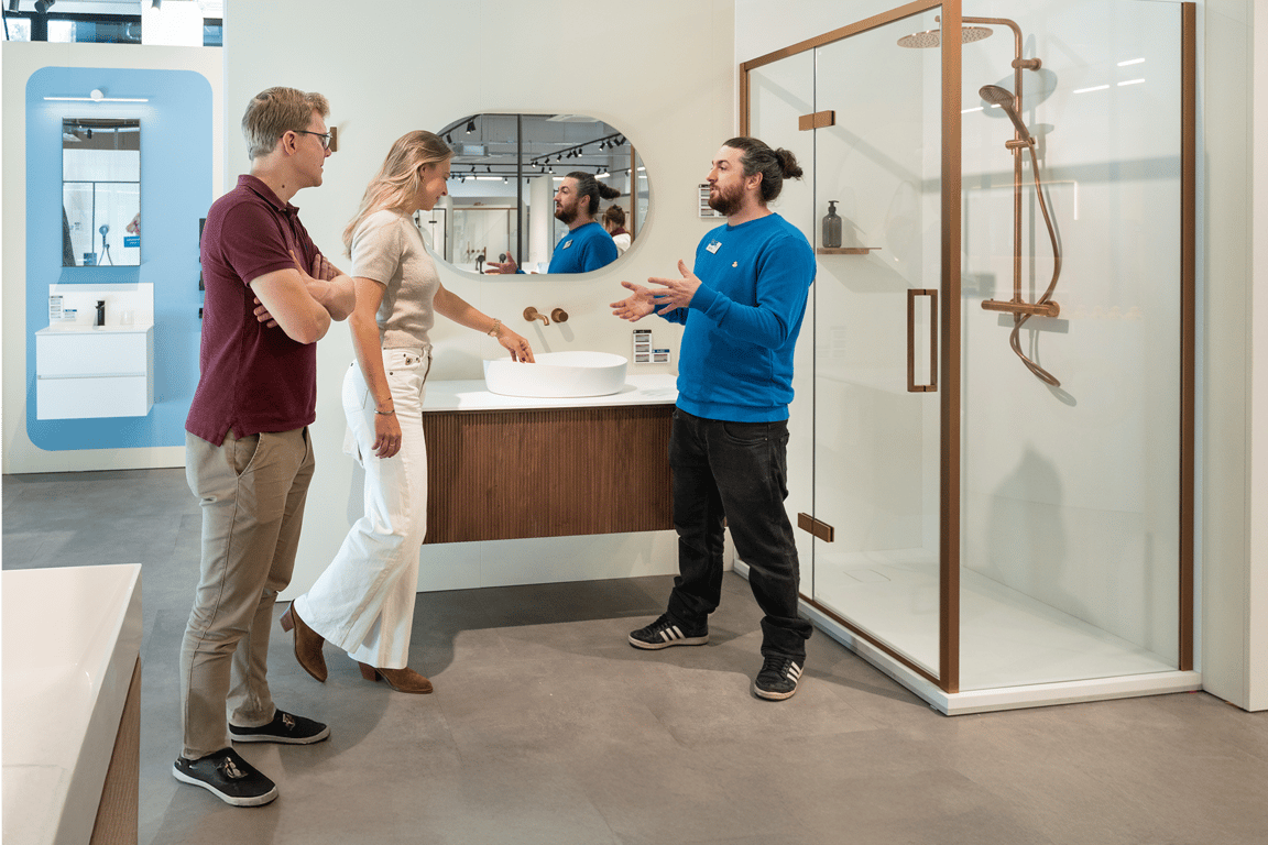 Een X²O badkamers-adviseur overhandigt brochures aan een lachend koppel tijdens een persoonlijk gesprek aan een ronde tafel in een moderne showroom met douches en kranen.