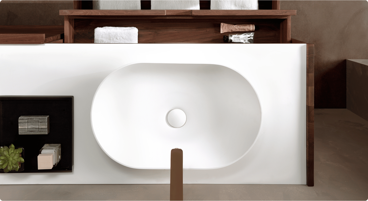 Bovenaanzicht van een minimalistische, witte wasbak ingebouwd in een meubel met houten accenten, onderdeel van een x²o badkamers interieur. Links op het werkblad staat een zwarte tray met decoratieve zepen en een klein vetplantje. Op de achtergrond zijn houten open vakken met handdoeken en verzorgingsproducten zichtbaar.
