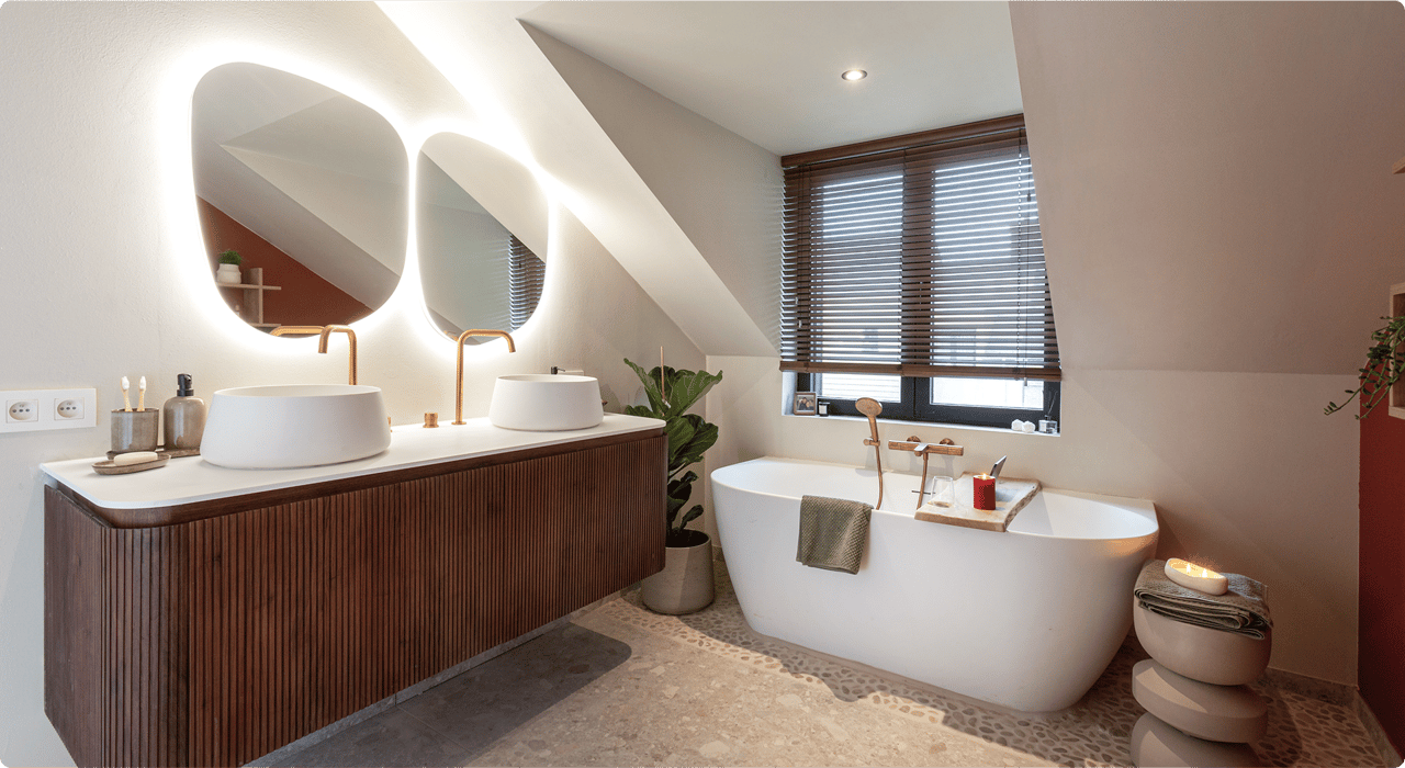 Moderne x²o badkamers inrichting onder een schuin dak met dubbele ronde spiegels, sfeerverlichting, twee vrijstaande waskommen met koperkleurige kranen, een vrijstaand bad voor het raam met houten jaloezieën en accessoires zoals kaarsen en planten.