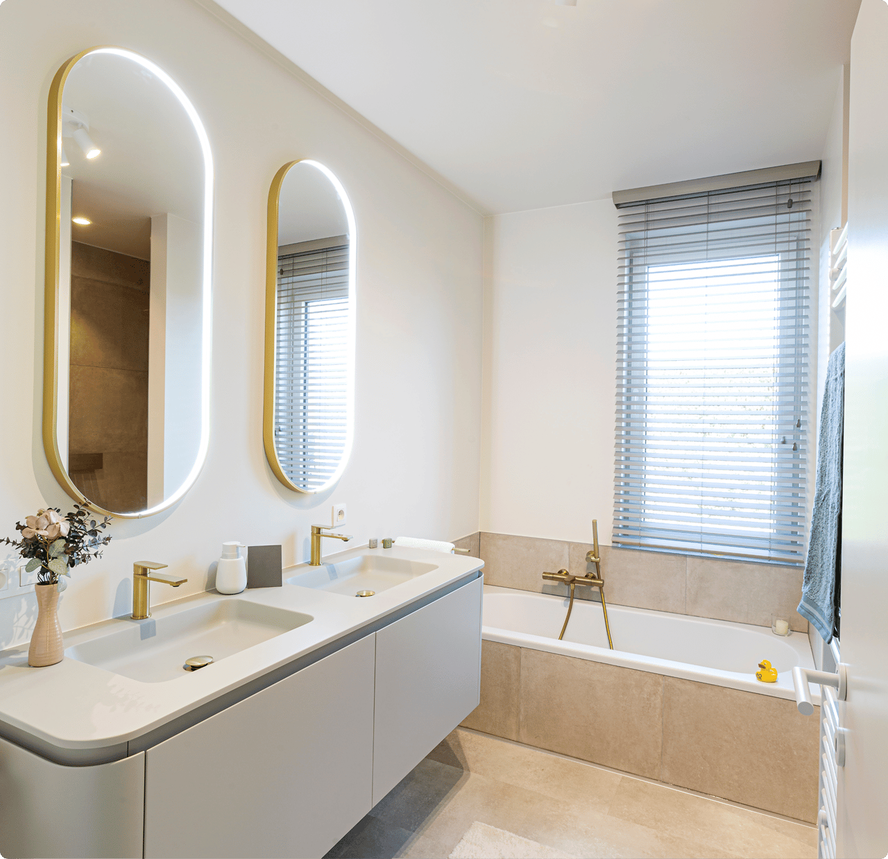 Lichte, frisse x²o badkamers met moderne indeling. Links een zwevend meubel met dubbele wastafel, goudkleurige kranen en twee verticale spiegels met ledverlichting. Rechts een ingebouwd ligbad met bijpassende kraan en een raam met jaloezieën zorgt voor natuurlijk licht.