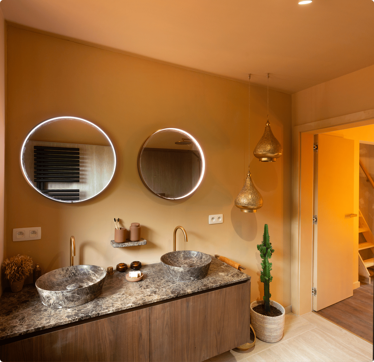 Warme, oosterse sfeer in deze x²o badkamers met aardetinten, twee ronde spiegels met geïntegreerde verlichting en een dubbel wastafelmeubel in hout met marmerlook werkblad. Op de achtergrond hangen decoratieve lantaarns en een cactus staat in een mand links van de deuropening.
