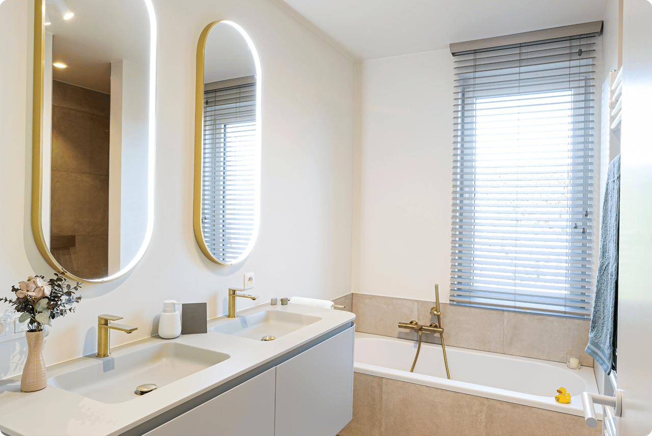 Lichte, frisse x²o badkamers met moderne indeling. Links een zwevend meubel met dubbele wastafel, goudkleurige kranen en twee verticale spiegels met ledverlichting. Rechts een ingebouwd ligbad met bijpassende kraan en een raam met jaloezieën zorgt voor natuurlijk licht.