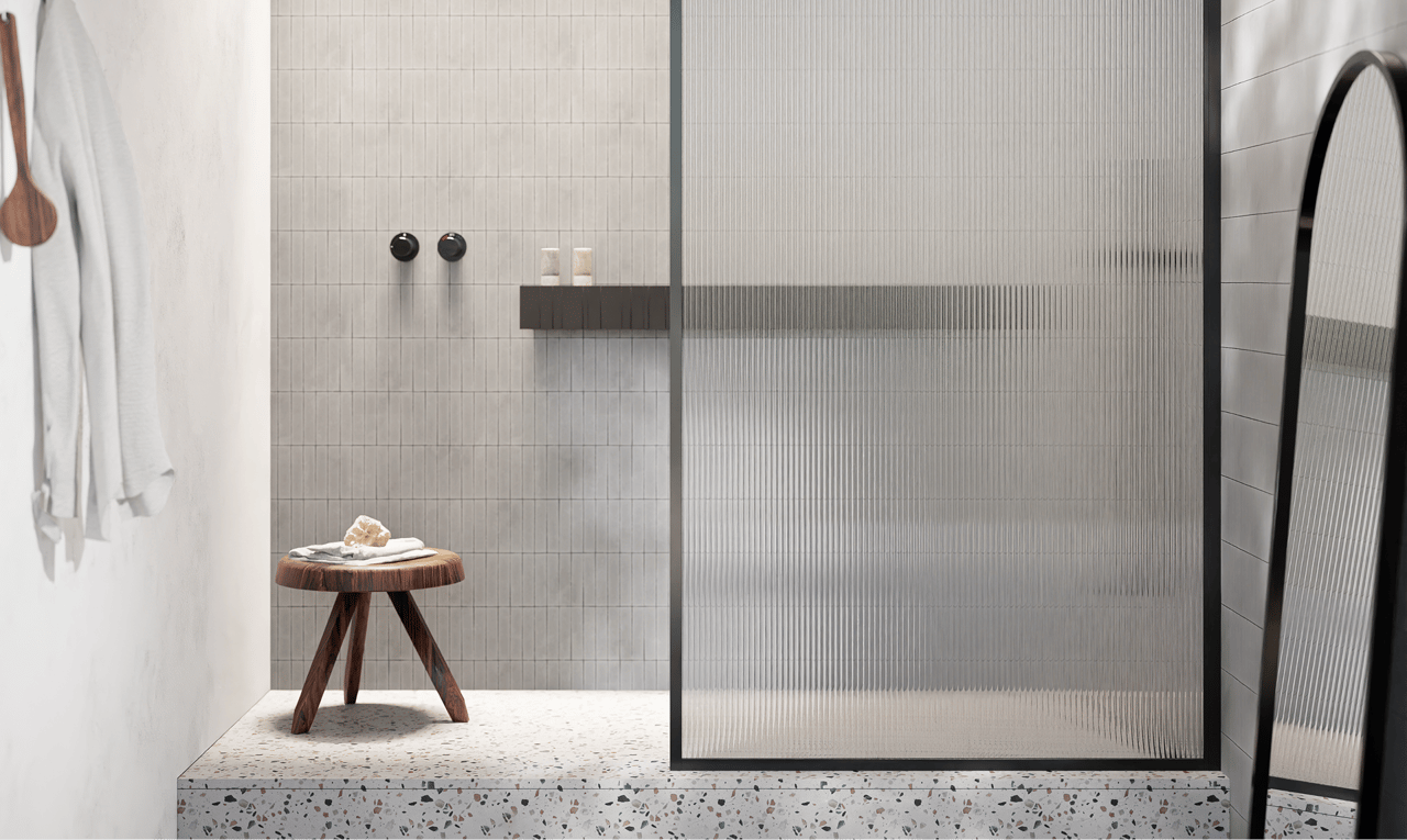 Moderne x²o badkamers inloopdouche met een verhoogd terrazzo-platform, fijne verticale wandtegels in lichtgrijs en een ribbelglas douchewand met zwart kader. Een houten krukje met een handdoek staat links in beeld, en rechts is een grote staande spiegel zichtbaar.