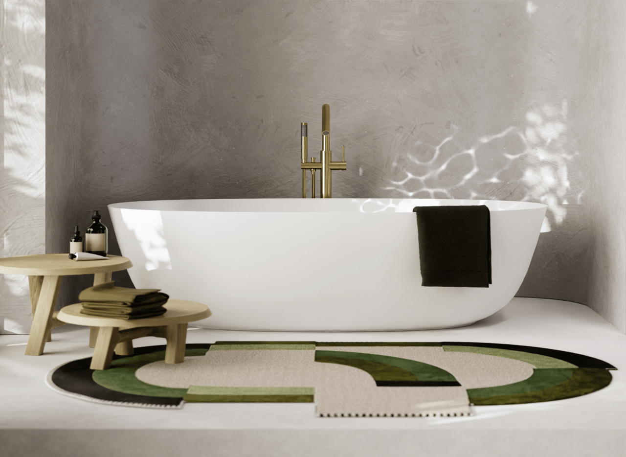 Lichte, moderne x²o badkamers setting met een vrijstaand wit bad, goudkleurige staande kraan en twee houten bijzettafels. Op de vloer ligt een opvallend rond vloerkleed in groen- en natuurtinten. Links zorgen grote ramen voor natuurlijk lichtinval, en rechts staat een ingekaderd kunstwerk.