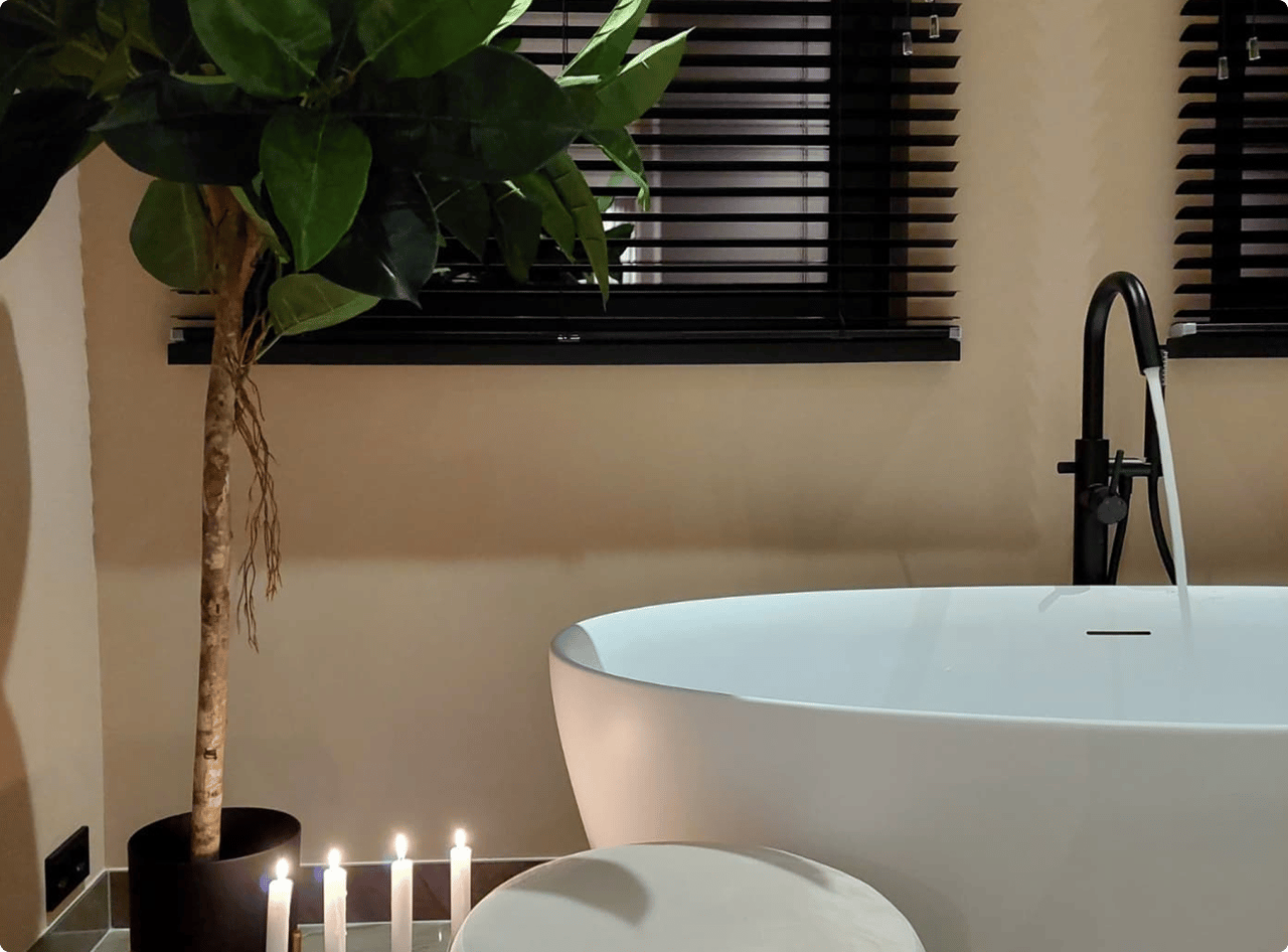 Sfeervolle x²o badkamers met een witte vrijstaande badkuip, zwarte staande kraan en een zacht verlichte setting. Zes brandende kaarsen en een grote groene kamerplant creëren een rustgevende sfeer. Op de achtergrond hangen zwarte jaloezieën voor het raam.