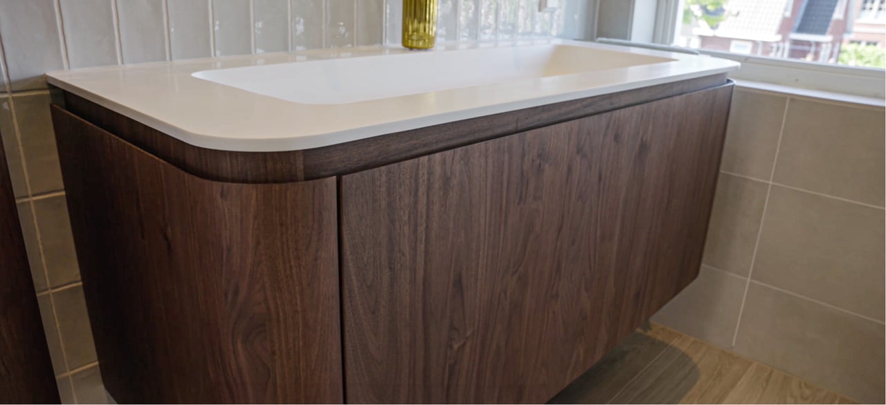 Nahaufnahme eines hängenden Holz-Badmöbels mit abgerundeten Ecken, darunter ein weißes Waschbecken. Im Hintergrund: geflieste Wand und Fenster. x²o Bad.