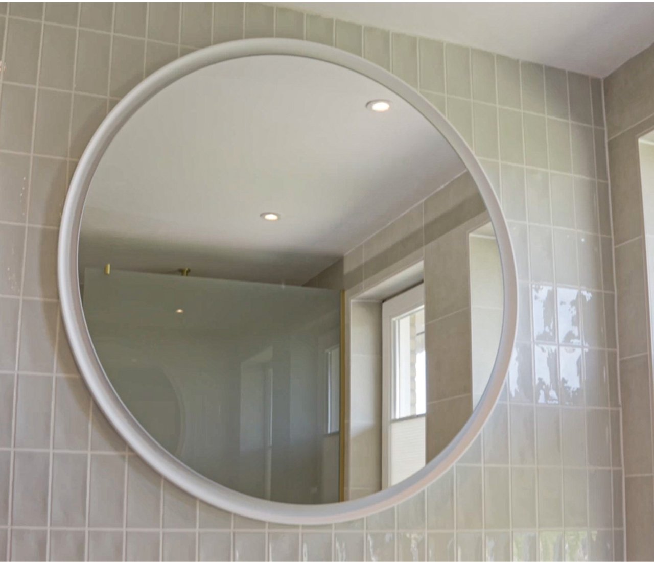 Runder Spiegel mit weißem Rahmen an einer Wand mit glänzenden rechteckigen Fliesen. Der Spiegel reflektiert Tageslicht und Einbauspots. x²o Bad.