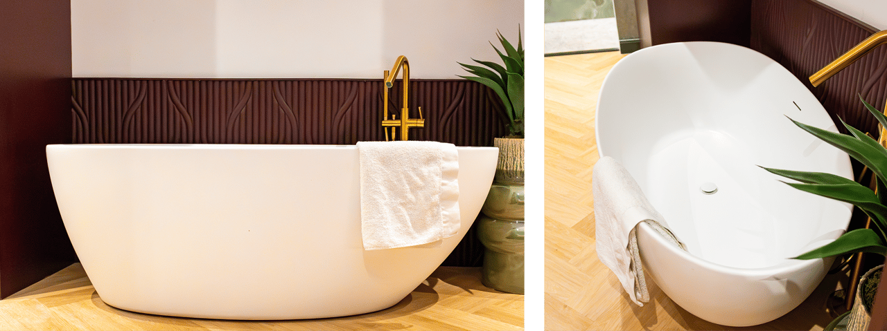 Auf diesem Foto siehst du eine stilvolle freistehende weiße Badewanne der x²o badkamers Ensuite mit goldener Armatur und einem weißen Handtuch darüber. Die Badewanne steht auf einem Fischgrät-Holzboden, daneben ein Blumentopf und ein dekoratives dunkelrotes Wandpaneel mit Relief.