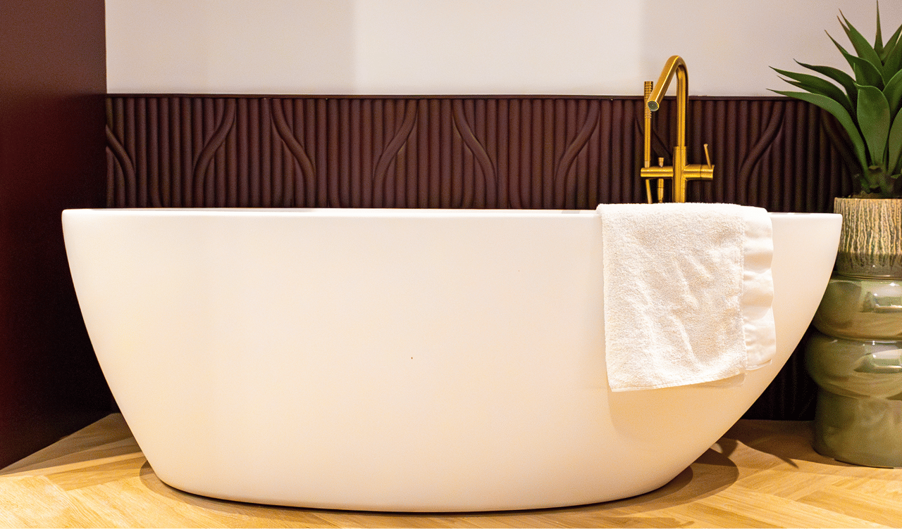 Auf diesem Foto siehst du eine stilvolle freistehende weiße Badewanne der x²o badkamers Ensuite mit goldener Armatur und einem weißen Handtuch darüber. Die Badewanne steht auf einem Fischgrät-Holzboden, daneben ein Blumentopf und ein dekoratives dunkelrotes Wandpaneel mit Relief.