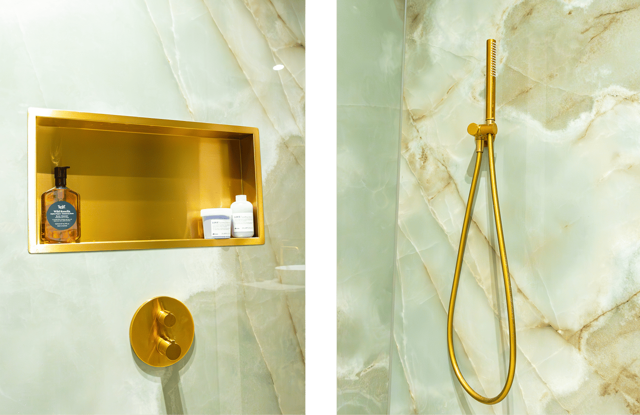 Auf diesem Foto siehst du eine Nahaufnahme der luxuriösen Dusche im x²o badkamers Ensuite-Badezimmer. Oben hängt ein großer runder Regenduschkopf in Gold. Daneben ist eine goldene Nische in die grün-marmorierte Duschwand eingelassen, darin stehen Pflegeprodukte wie ein Pumpspender und kleine Tiegel. Außerdem ist eine goldene Handbrause mit flexiblem Schlauch an der Marmorwand befestigt.