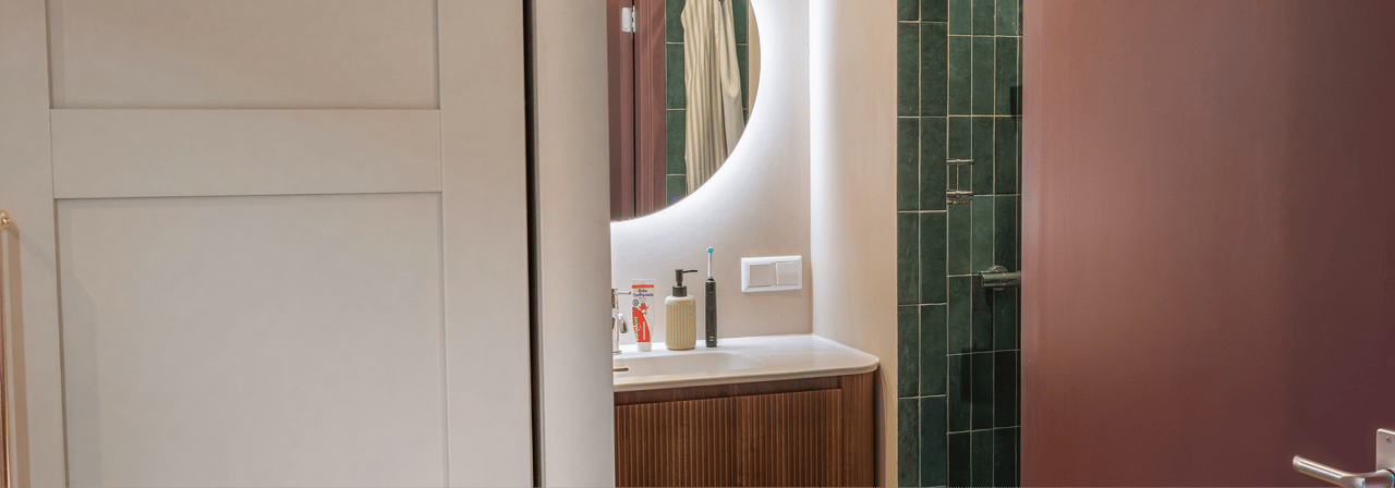 Gedeeltelijk zicht op een badkamer x²o badkamers met een halfopen deur die uitkijkt op een houten geribbeld wastafelmeubel met witte waskom, chromen kraan, ronde verlichte spiegel en rechts een douche met groene tegels en glazen wand