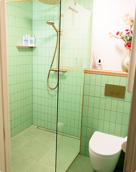 In diesem x²o Badezimmer siehst du eine begehbare Dusche mit goldener Regendusche und Handbrause, grüne Wandfliesen, eine Glasabtrennung, ein wandhängendes WC mit goldener Drückerplatte und ein Holzbrett mit Deko.