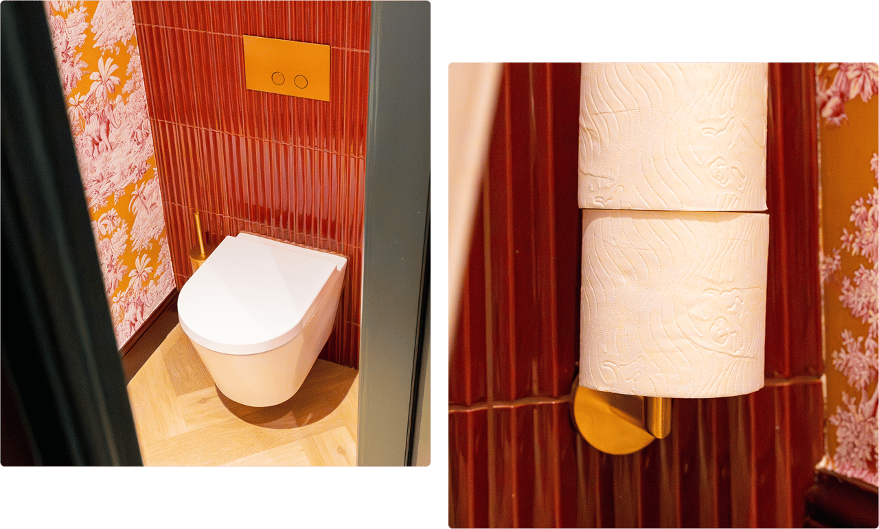 Ein weißes Hänge-WC von x²o badkamers steht vor einer Wand mit roten vertikalen Fliesen und orange-rosa Tapete mit tropischen Motiven, dazu eine goldene WC-Bürste und eine eingebaute Spülplatte.