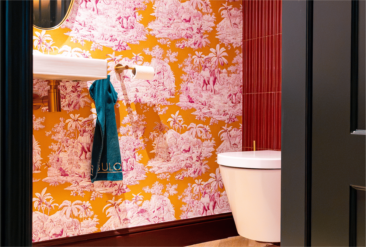 Goldener Toilettenpapierhalter im Stil von x²o Badezimmer mit zwei Rollen, vor roten Wandfliesen und rosafarbener Tapete.