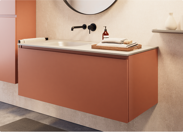 x²o badkamers met warme kleuraccenten dankzij een matte oranje wastafelkast, zwarte kraan, ronde spiegel en gestileerde accessoires op een beige wand.