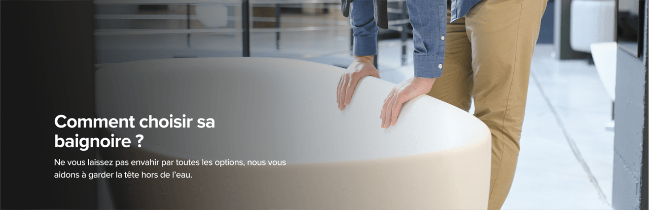 Sur cette image de x²o badkamers, un homme en pantalon beige et chemise bleue examine une grande baignoire design van x²o badkamers in een showroom met een industriële vloer en andere badmodellen op de achtergrond.