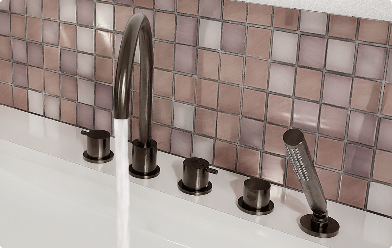 Op deze foto van x²o badkamers zie je een op de badrand gemonteerde badkraan in bronskleur, met een hoge gebogen uitloop waar water uit stroomt, vier ronde bedieningsknoppen op een rij en een cilindrische handdouche, alles geplaatst voor een wand met glanzende vierkante tegeltjes in roze en paarse tinten.