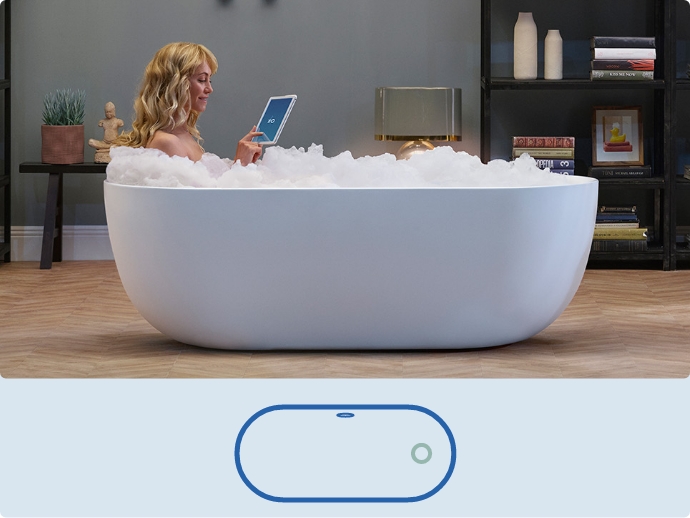 Witte vrijstaande badkuip gevuld met schuim, een vrouw leest ontspannen een tablet.