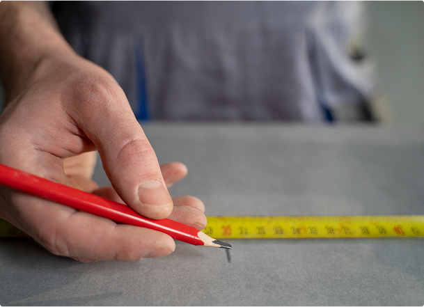 Een close-up van een hand die met een rood potlood een maat aanduidt op een grijze ondergrond, naast een gele meetlint. Deze nauwkeurige werkwijze maakt deel uit van de voorbereiding in een x²o badkamers renovatie of installatie.