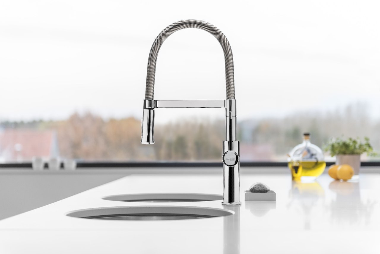 Chroomkleurige kraan met flexibele slang op wit aanrecht met twee gootstenen, op de achtergrond olijfolie en citroenen.