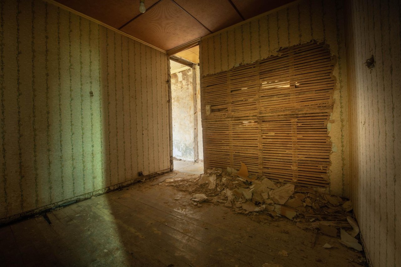 Verlassene Zimmer mit beschädigter Wand, abgenutztem Tapeten und Schutt auf dem Boden. Das Licht fällt durch das Fenster