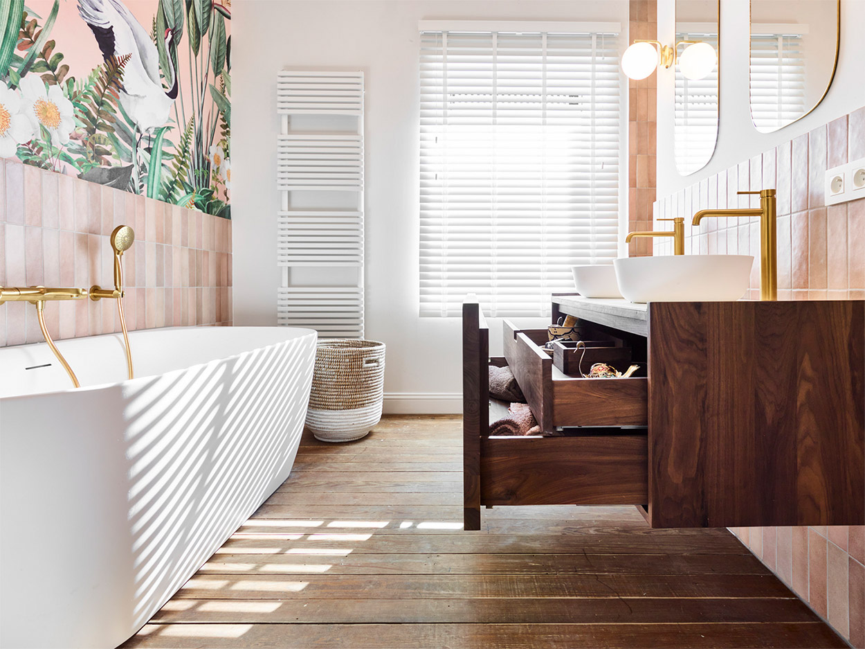 Stijlvolle badkamer met een witte badkuip, gouden kranen, tropische muurschildering, houten wastafelmeubel en opberglade