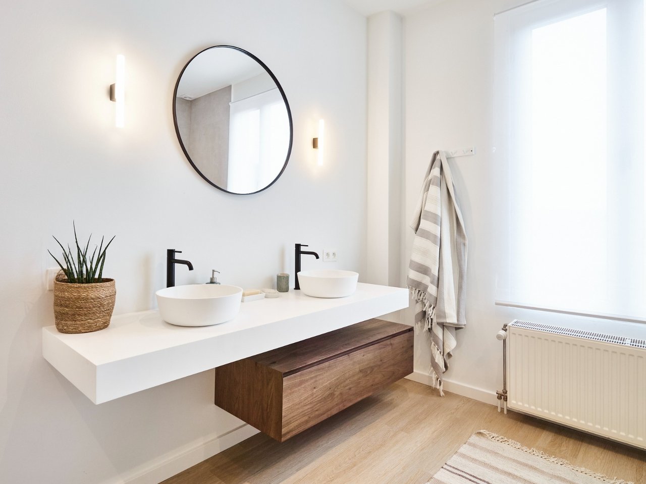 Salle de bain moderne avec deux lavabos blancs, robinets noirs et un miroir rond au-dessus d'un meuble en bois.