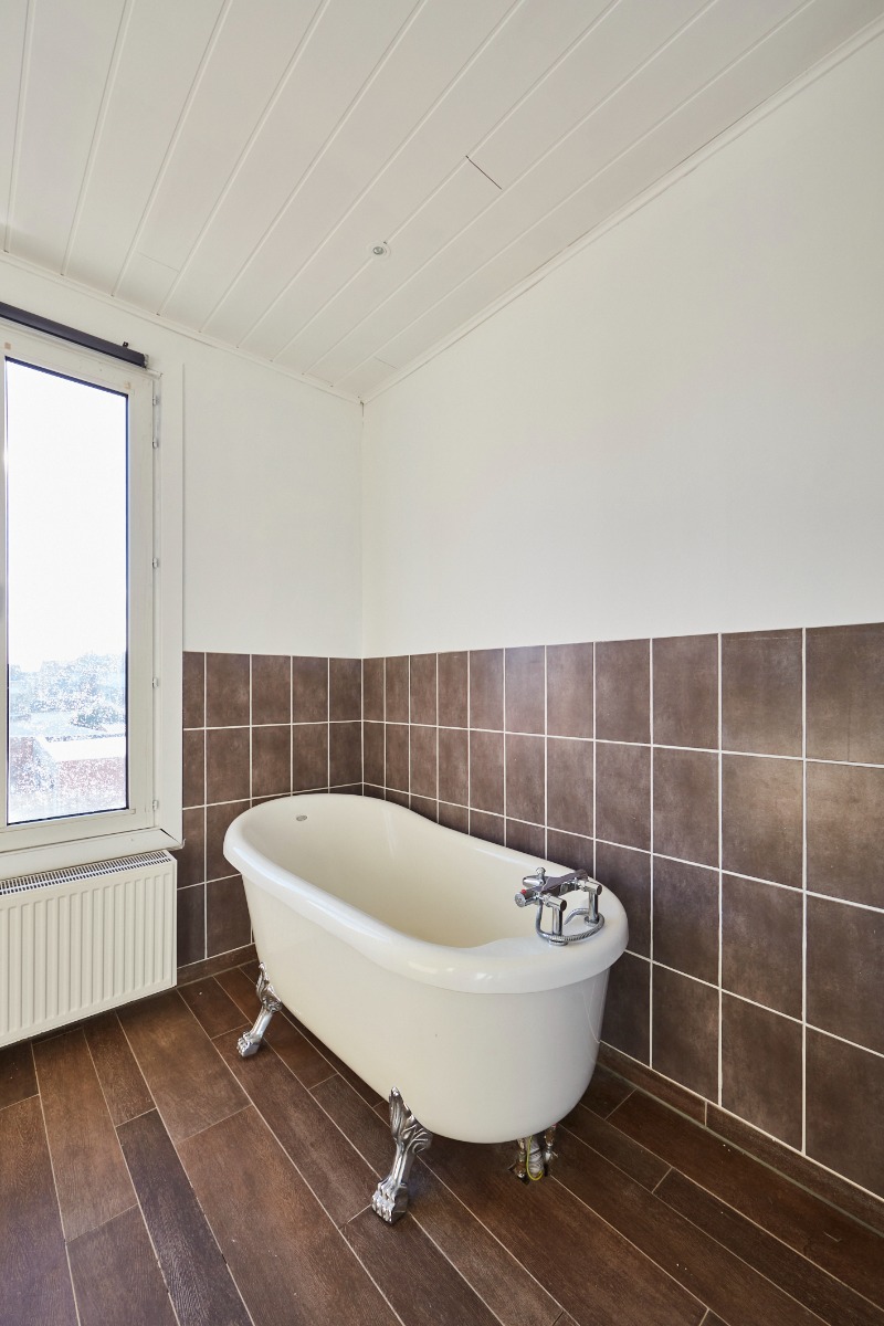 Baignoire sur pied avec des pieds classiques, contre un mur carrelé marron et une fenêtre à côté.