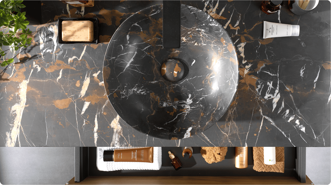 Close-up van een dubbele wastafelopstelling van x²o badkamers met twee ronde waskommen in donker marmerpatroon, zwarte kranen en een houten meubel met verticale groeven. Op het werkblad staan verzorgingsproducten, een tandenborstel en een borstel. Op de achtergrond hangen twee spiegels met geïntegreerde verlichting en daarachter een douchecabine met glazen wand en zwarte accenten.