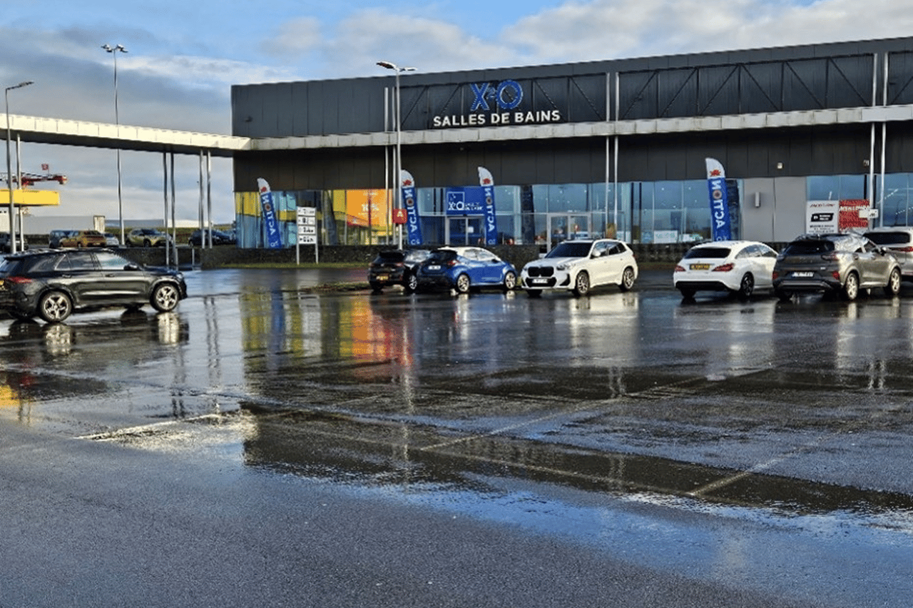 Voorzijde van een moderne winkel van XO Badkamers met een natte parkeerplaats en verschillende geparkeerde auto's op een bewolkte dag.