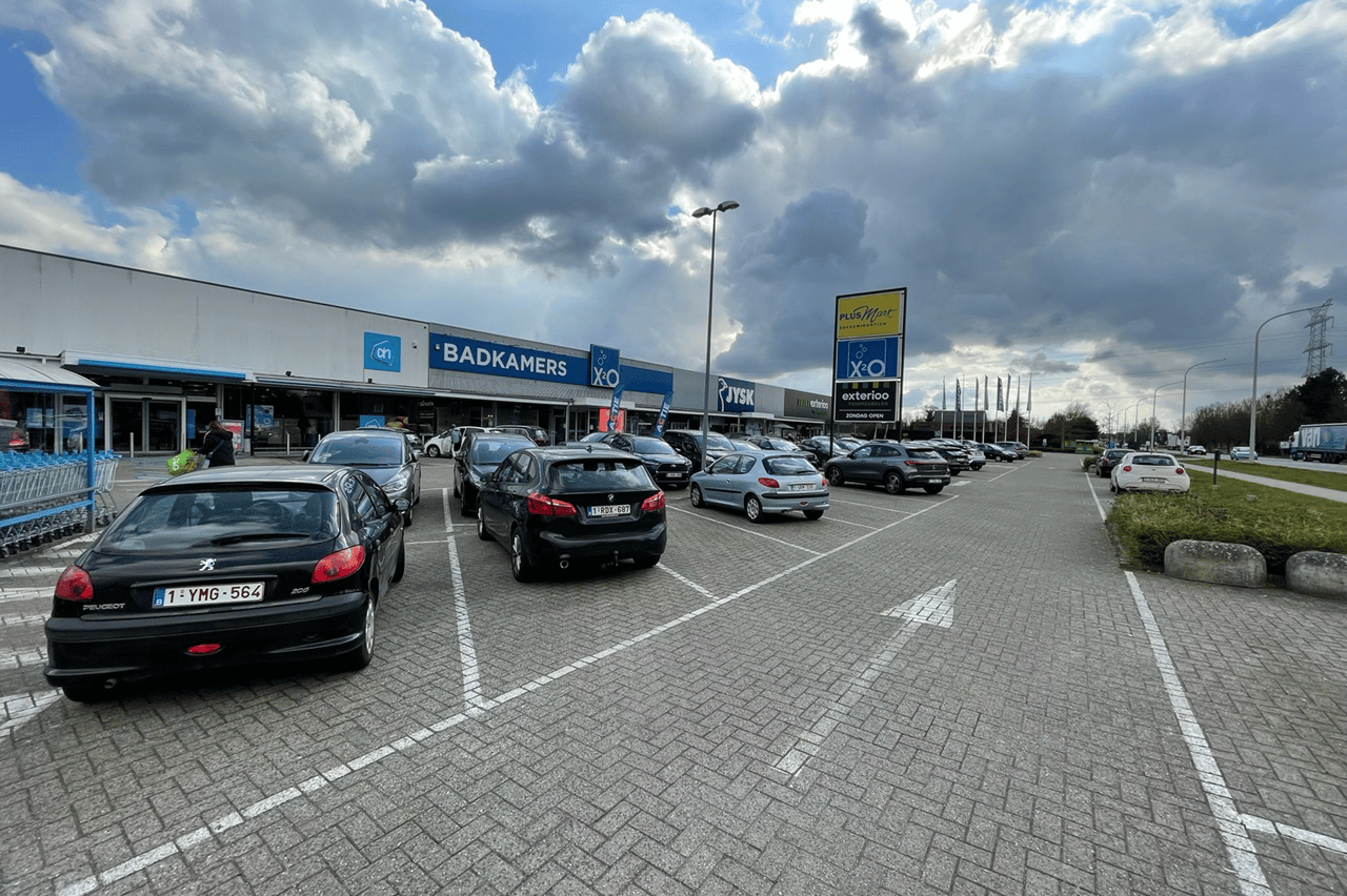 Drukke parkeerplaats voor een winkelcentrum met onder andere XO Badkamers, onder een deels bewolkte hemel met zonnestralen door de wolken.