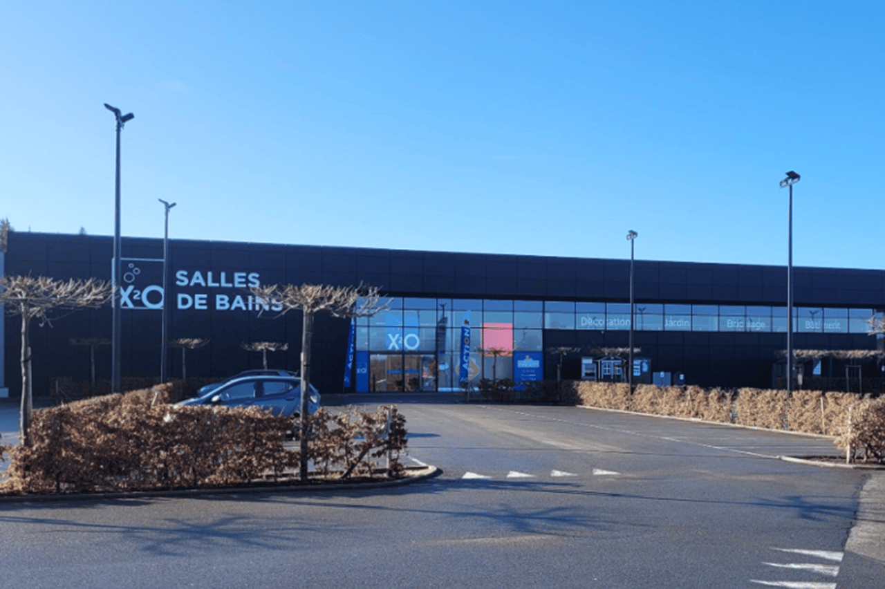 Voorzijde van een moderne XO Badkamers-winkel met zwarte gevel, een lege parkeerplaats en strak blauwe lucht op een zonnige dag.