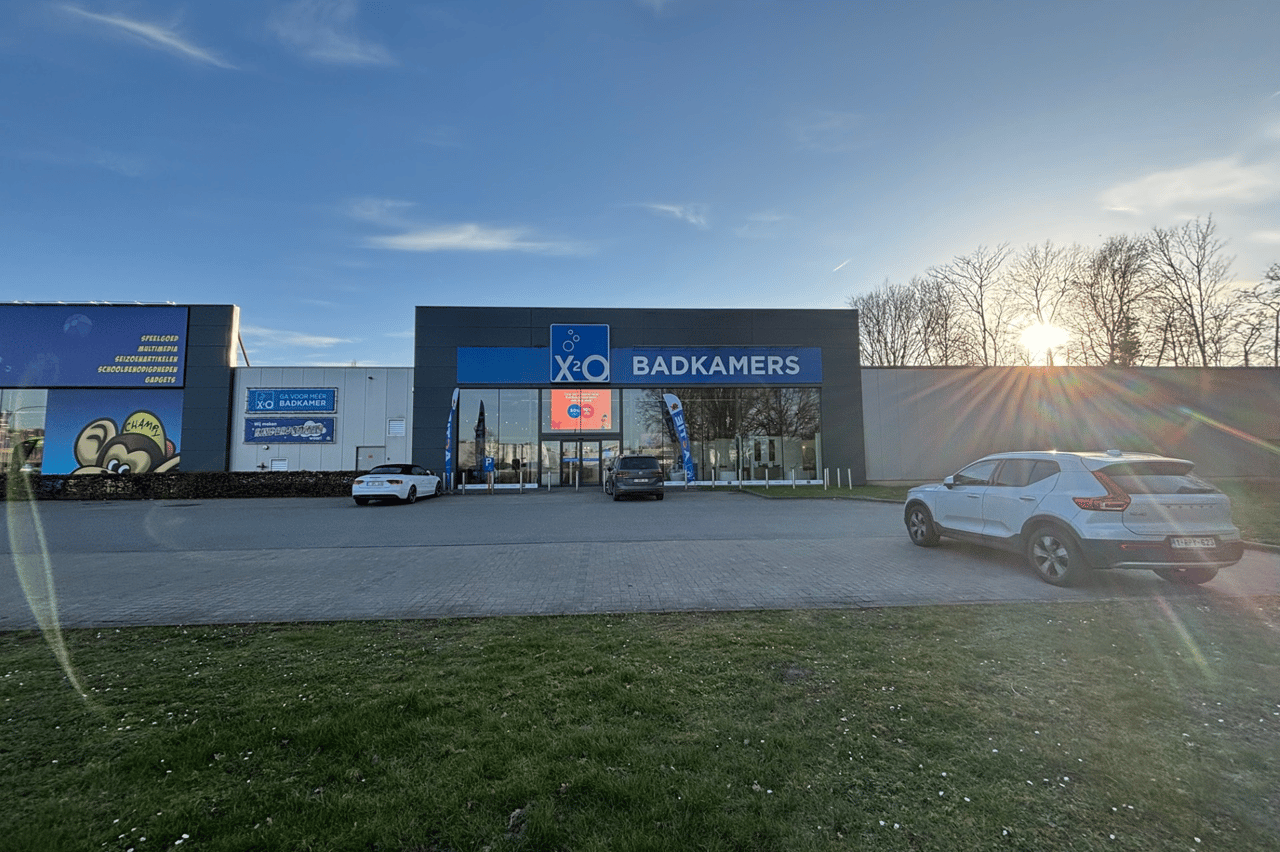 X2O Badkamers-winkel met glazen gevel en enkele geparkeerde auto's op een open terrein, met de zon die ondergaat achter een rij bomen.