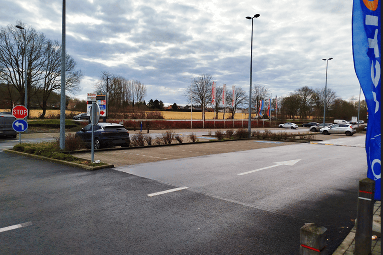 Uitrit van een parkeerterrein met een STOP-bord, enkele geparkeerde en passerende auto's, vlaggenmasten in de verte en een bewolkte lucht.