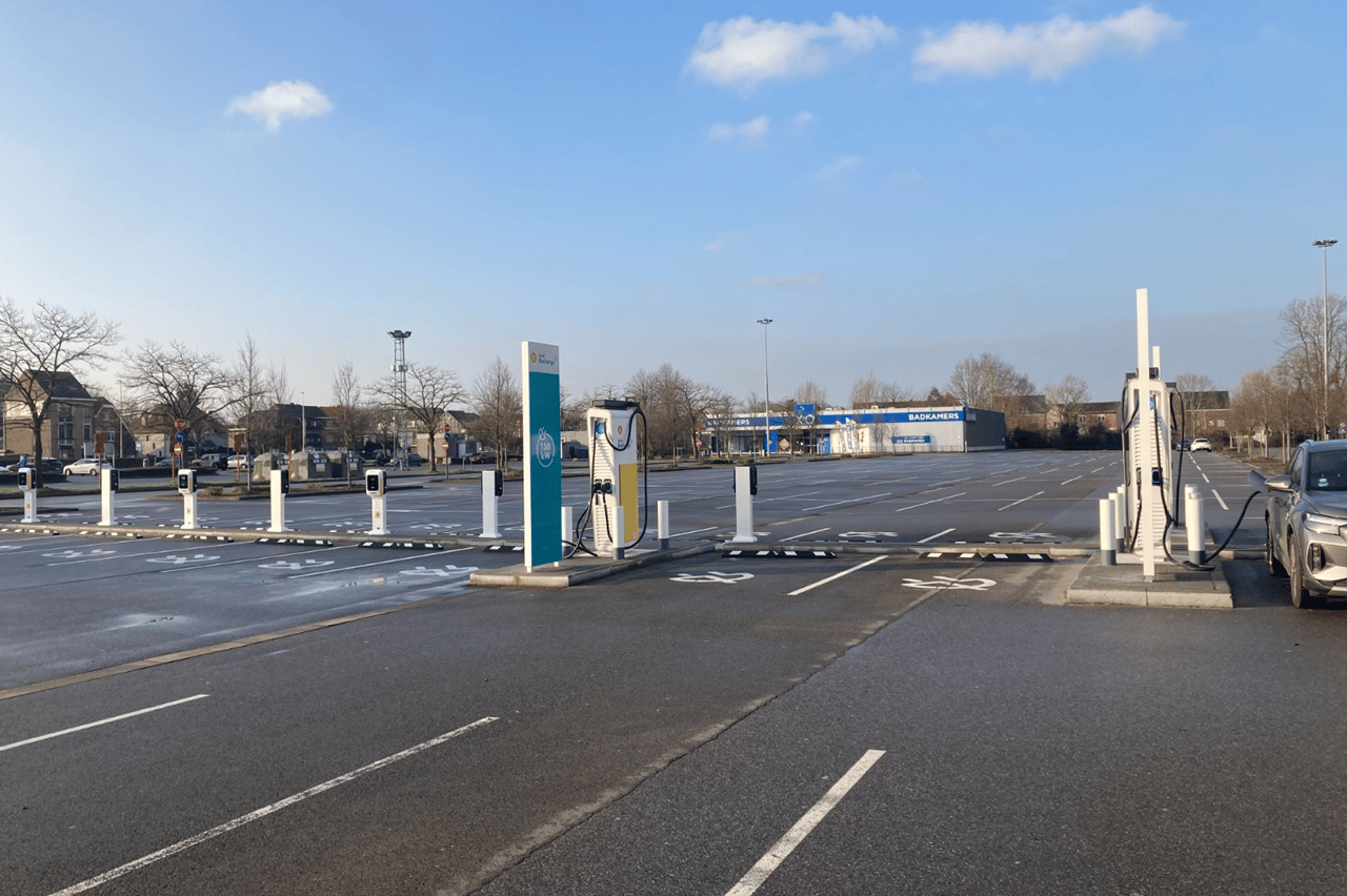 Grote, grotendeels lege parkeerplaats met meerdere elektrische laadpalen en op de achtergrond een XO Badkamers-winkel onder een heldere blauwe lucht.
