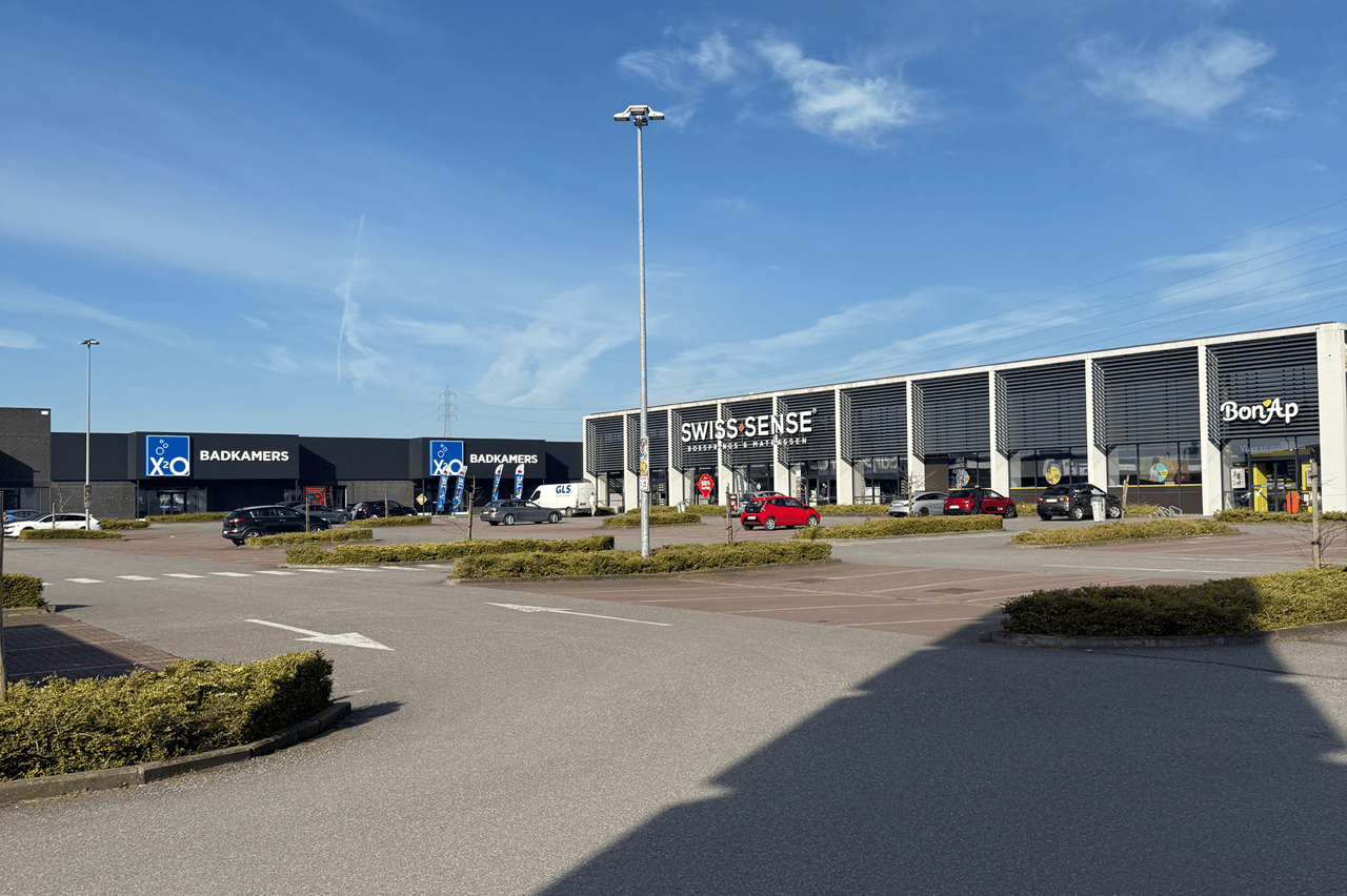 Winkelcomplex met onder andere X2O Badkamers, Swiss Sense en Bon’Ap, met parkeerplaatsen en een brede oprijlaan onder een helderblauwe hemel.