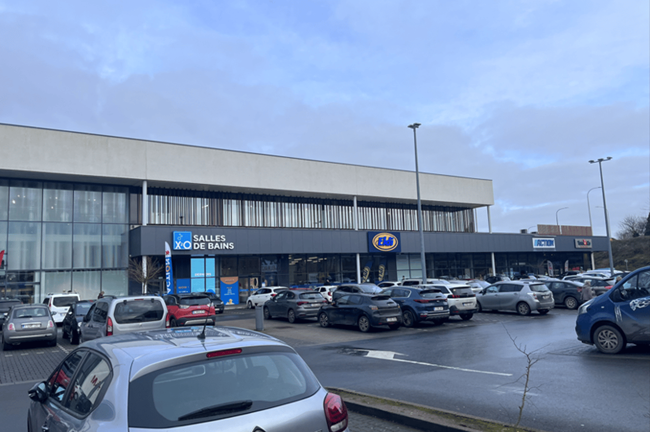 Winkelcentrum met X2O Badkamers, JBC en Action, voor een drukke parkeerplaats op een bewolkte dag.