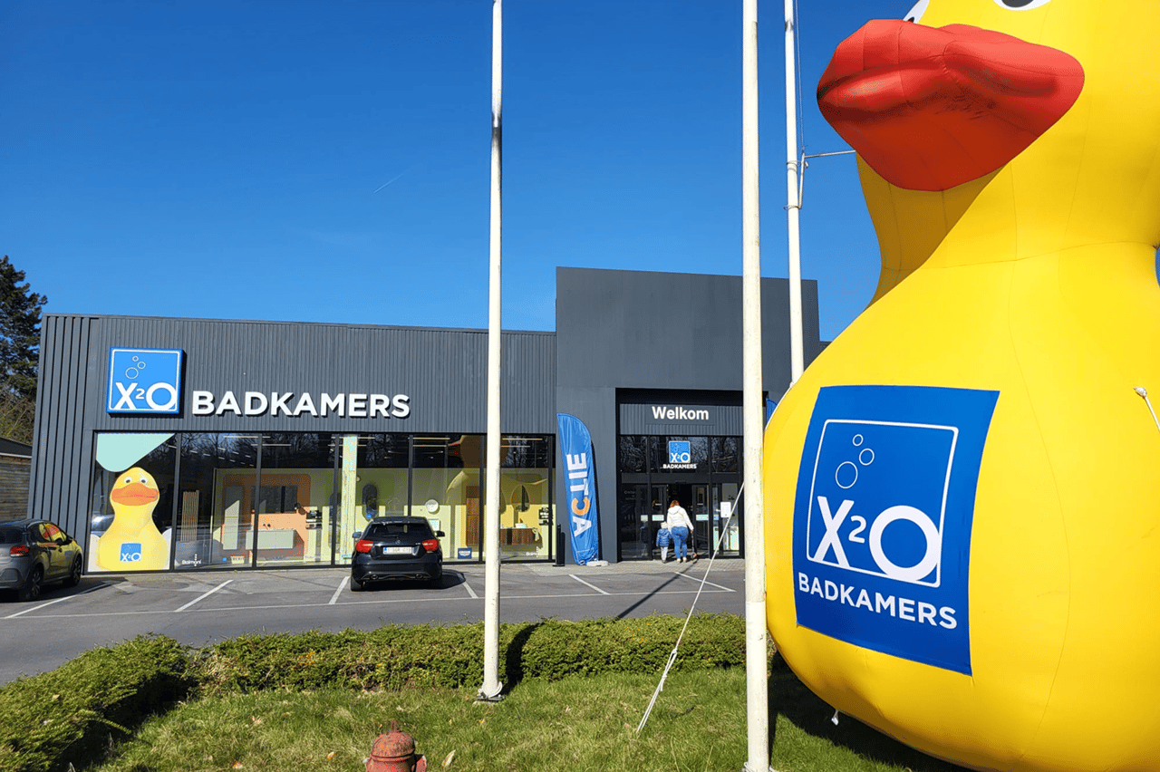 X2O Badkamers-winkel met grote opblaasbare gele badeend met logo op de voorgrond, onder een stralend blauwe hemel.