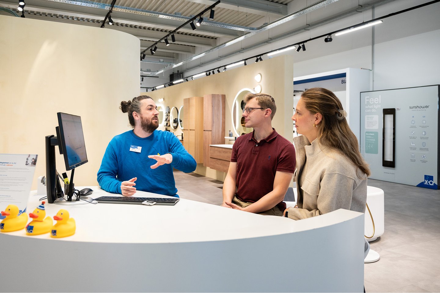 In een showroom van x²o badkamers geeft een medewerker uitleg aan een koppel aan een ronde balie met computer. Op de balie staan gele badeendjes als decoratie. Op de achtergrond zijn verschillende badkameropstellingen zichtbaar met spiegels, meubels en verlichting in een moderne winkelomgeving.