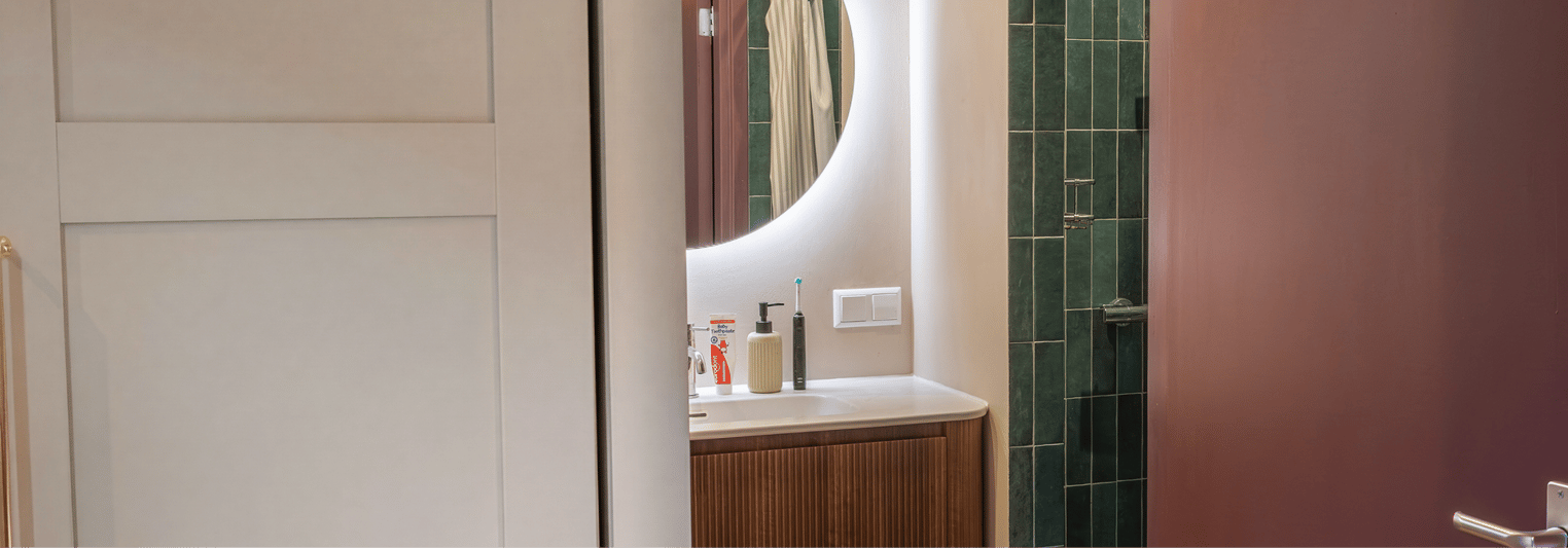 Gedeeltelijk zicht op een badkamer x²o badkamers met een halfopen deur die uitkijkt op een houten geribbeld wastafelmeubel met witte waskom, chromen kraan, ronde verlichte spiegel en rechts een douche met groene tegels en glazen wand