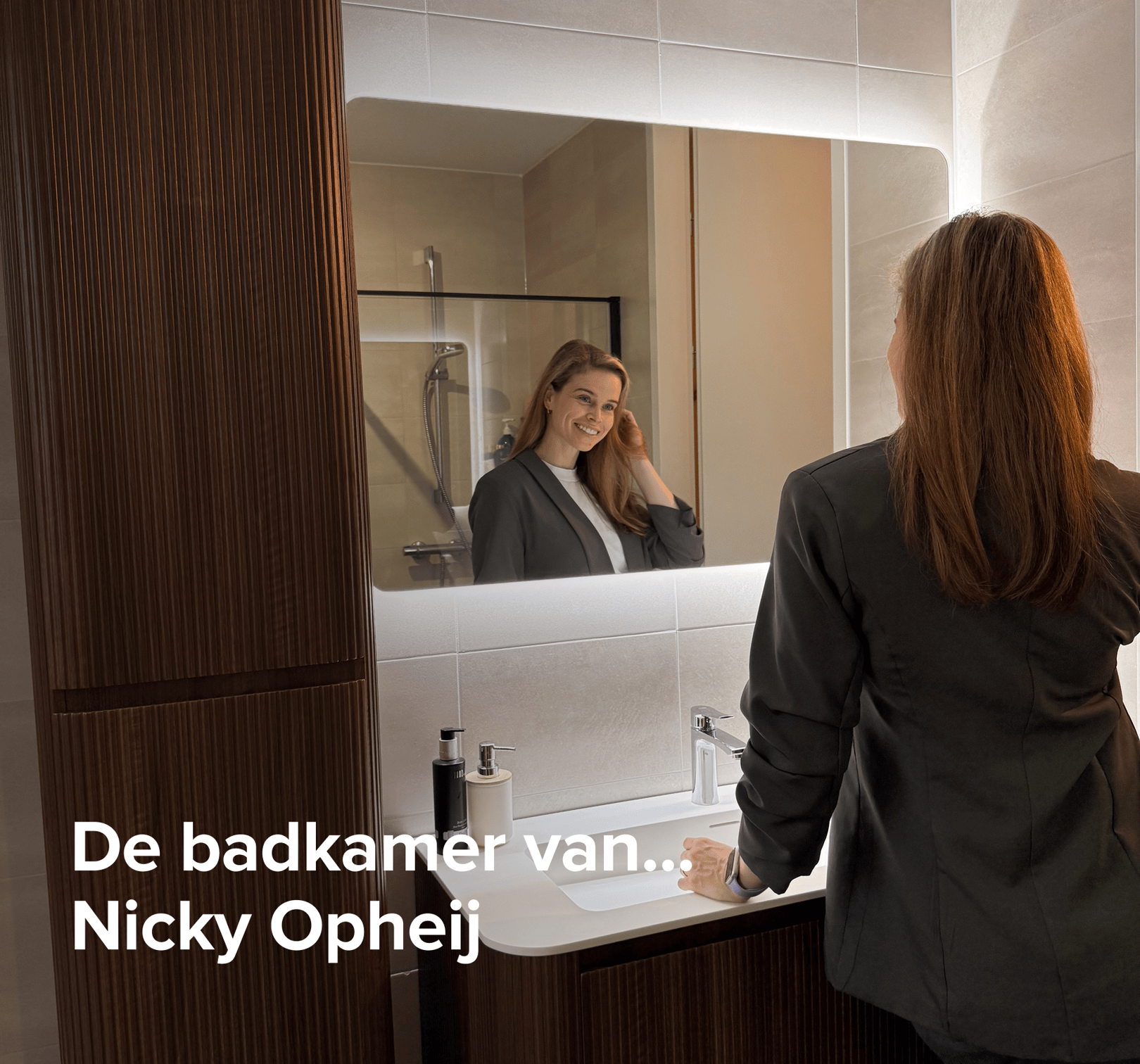 In deze inspirerende scène van x²o badkamers zie je een stijlvolle badkamer met een houten meubel en een verlichte spiegel. Een vrouw kijkt glimlachend naar zichzelf in de spiegel terwijl ze haar haar aanraakt. Op de afbeelding staat de tekst: “De badkamer van… Nicky Opheij”.